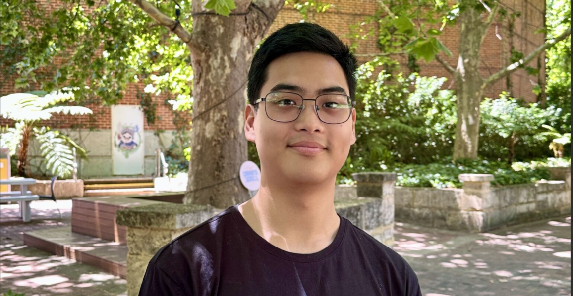 Lawrence stands smiling in front of trees. He wears a black tshirt and glasses.
